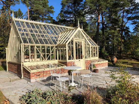 Vackert växthus, KEW Crane, byggt i Sverige med höga tallar i bakgrunden. Sittgrupp i solen.